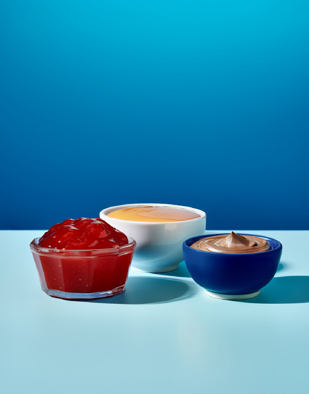 Schmuckbild Trockenwaren . Drei Schüsseln mit Marmelade, Pudding und Nussnougatcreme vor blauem Hintergrund